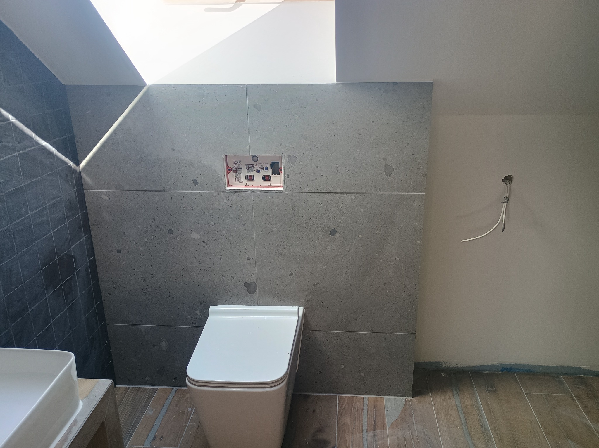 Nowoczesna łazienka w trakcie wykańczania: wisząca toaleta, szare płytki imitujące kamień, drewnopodobne panele podłogowe i okno dachowe wpadające do wnętrza