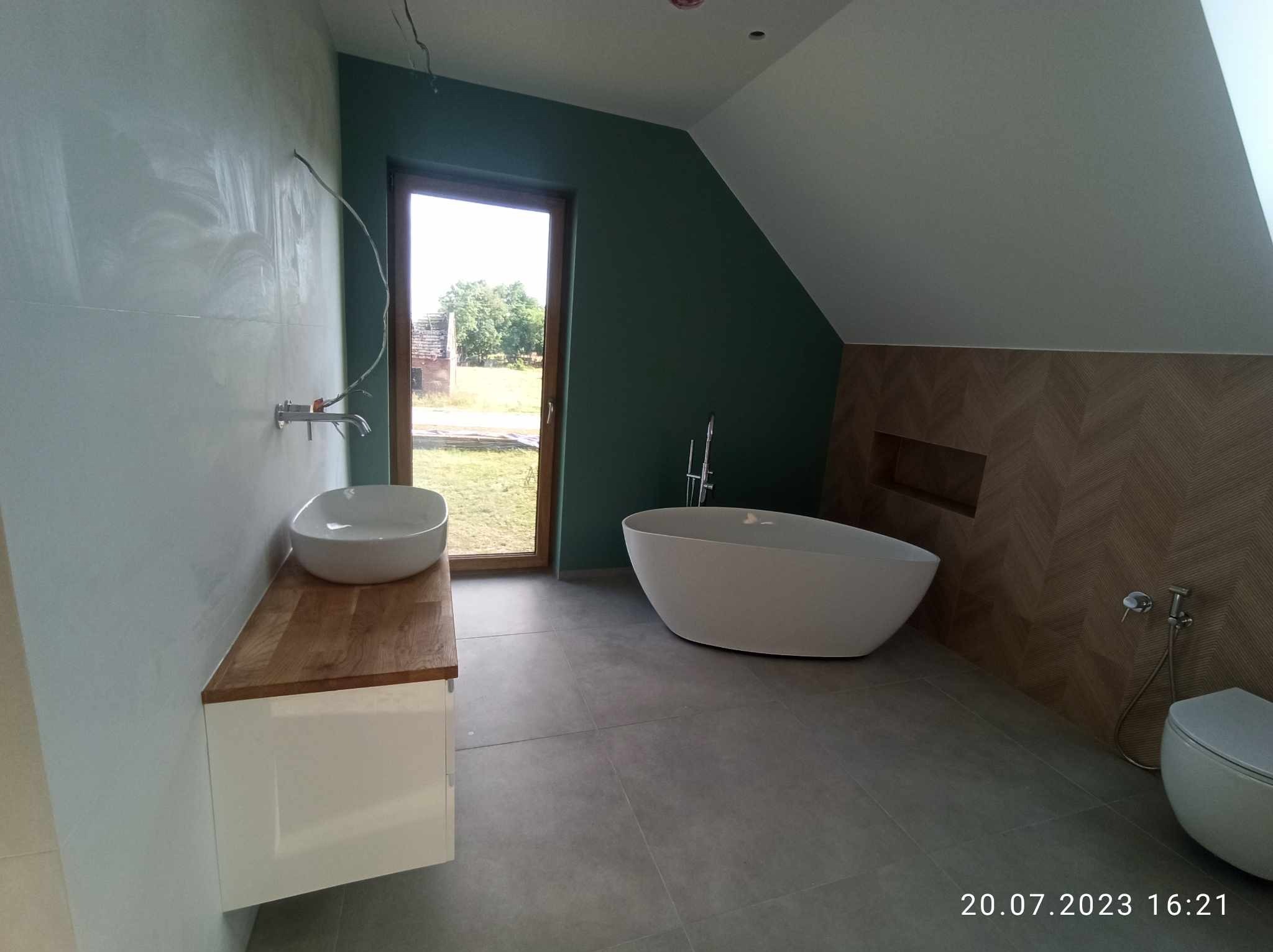 Nowoczesna łazienka na poddaszu z wolnostojącą wanną, umywalką nablatową na drewnianym blacie, szarą podłogą i zieloną ścianą z widokiem na ogród przez okno.