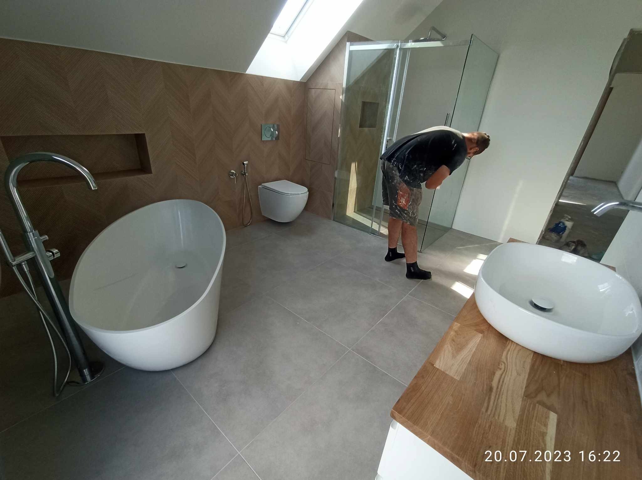 Nowoczesna łazienka na poddaszu z wolnostojącą wanną, umywalką nablatową na drewnianym blacie i kabiną prysznicową typu walk-in. Widoczny pracownik w ubraniach roboczych.