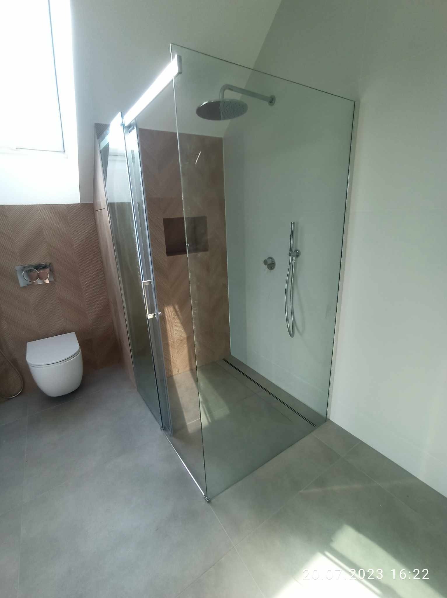 Nowoczesna łazienka z kabiną prysznicową typu walk-in, wykończona płytkami imitującymi drewno i szarą podłogą. Widoczny biały sedes podwieszany oraz deszczownica i bateria prysznicowa.