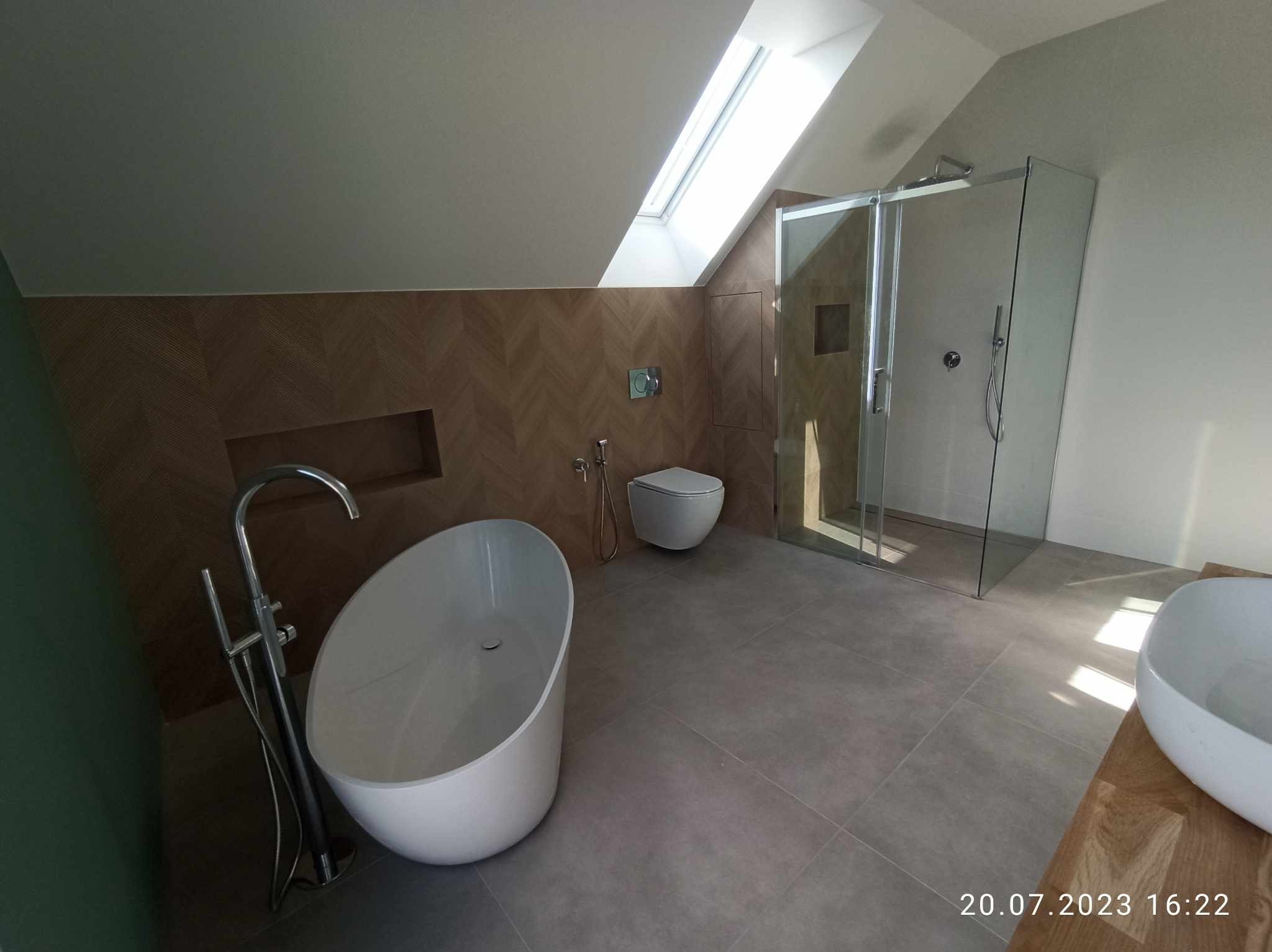 Nowoczesna łazienka na poddaszu ze skośnym sufitem i oknem dachowym, wolnostojąca wanna z chromowaną baterią, kabina prysznicowa ze szklanymi drzwiami, wisząca toaleta i drewniana okładzina ściany...