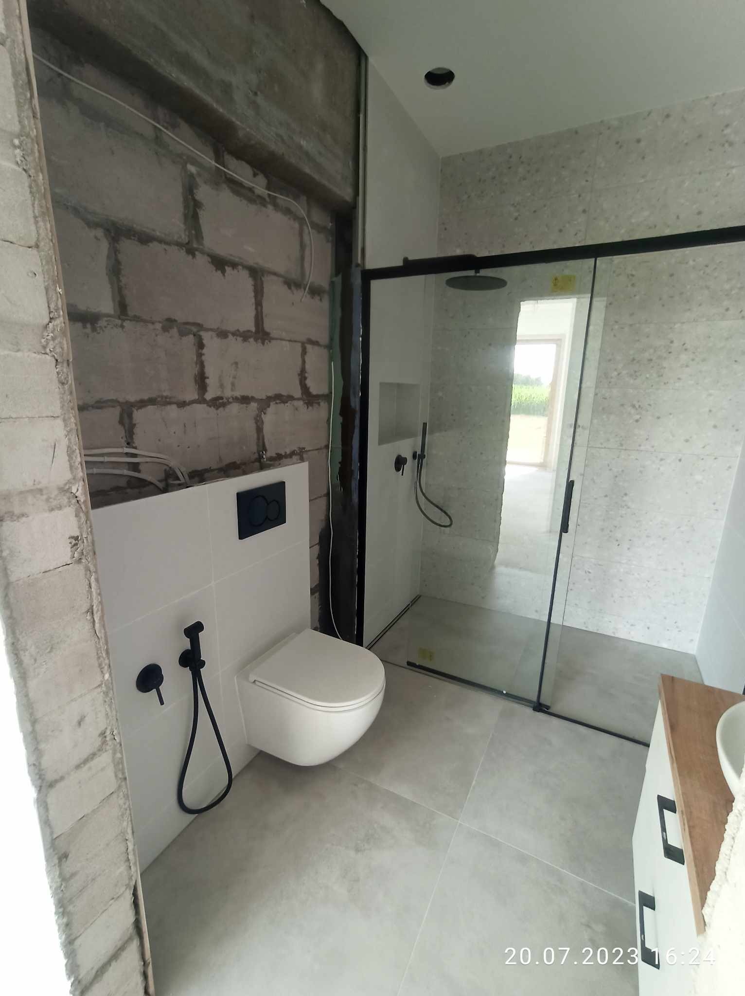 Wnętrze łazienki w trakcie remontu: surowy mur z instalacjami obok wykończonej części z białą ceramiką, czarną armaturą i kabiną prysznicową z czarnymi profilami.