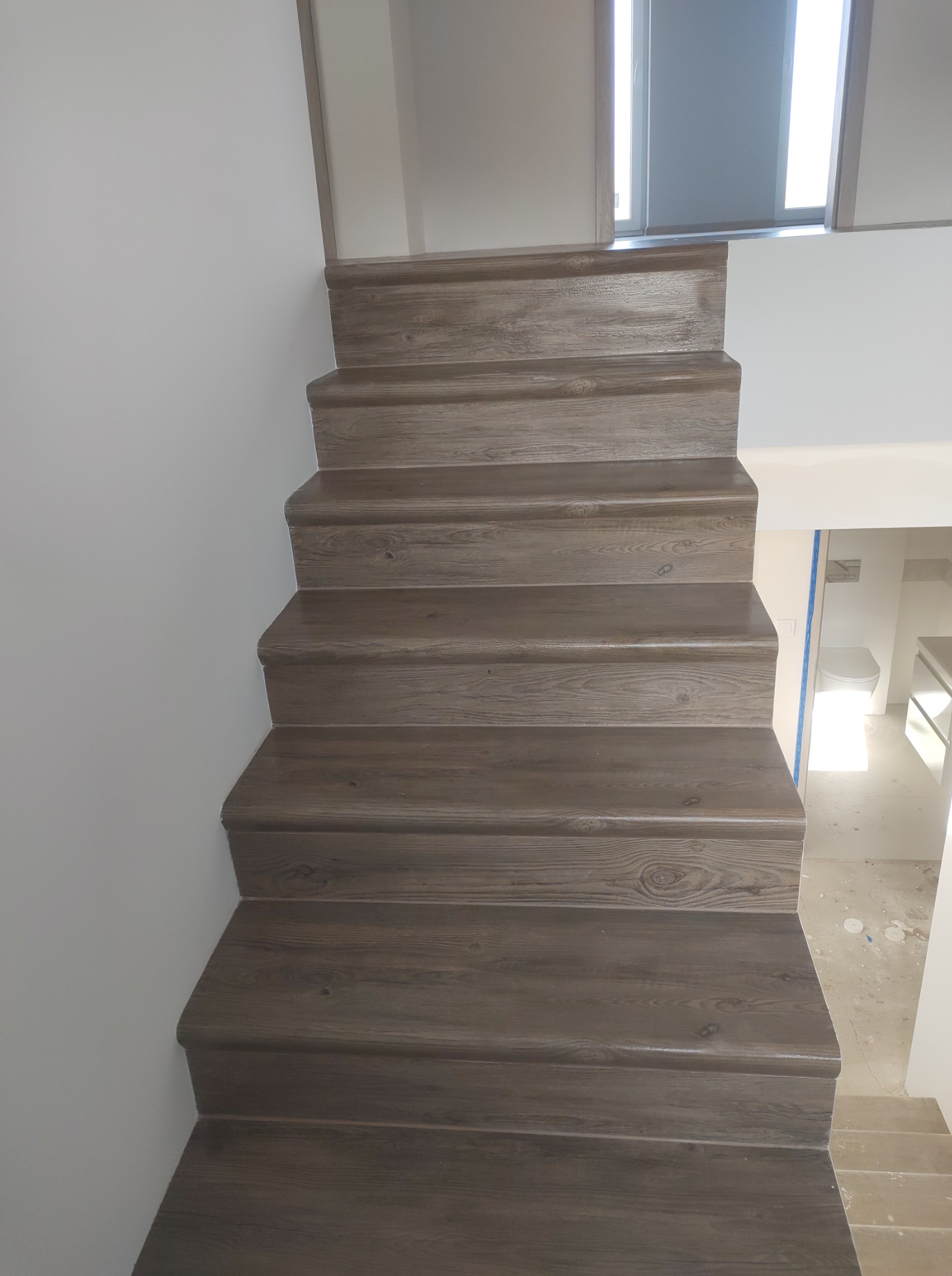 Drewniane schody z widocznym wykończeniem i fragmentem łazienki w tle, z toaletą i umywalką, ukazujące prace remontowe.