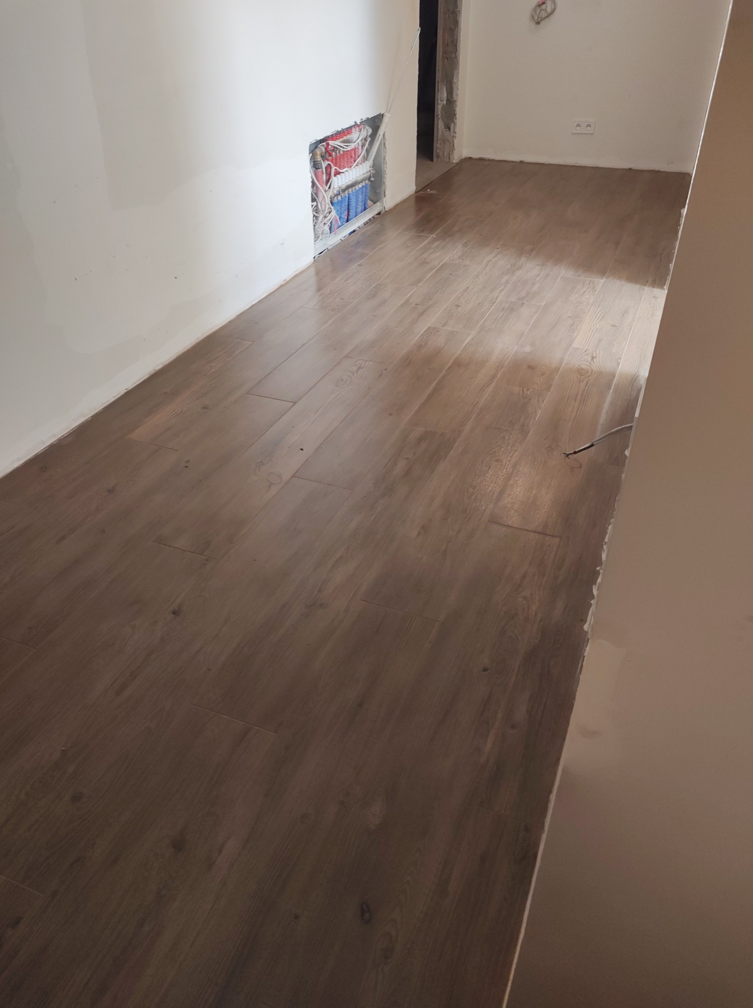 Ułożona podłoga z paneli imitujących drewno w pomieszczeniu w trakcie remontu, widoczne kable w ścianie i surowy stan wykończenia.