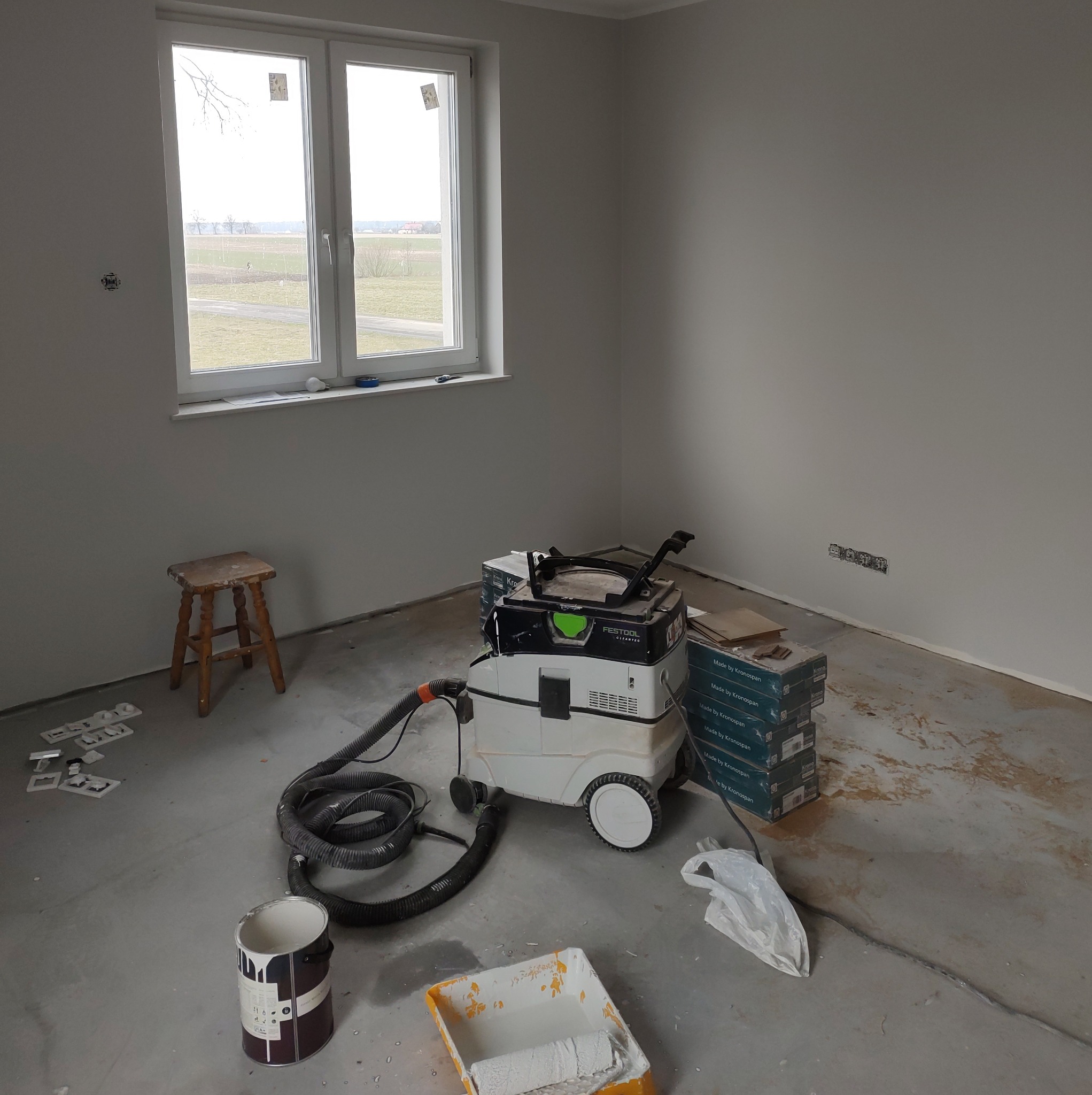 Przygotowania do malowania ścian w nowym budynku: wiadro z farbą, kuweta z wałkiem, odkurzacz przemysłowy Festool i paczki paneli podłogowych Kronospan stoją na surowym betonowym podłożu przy...