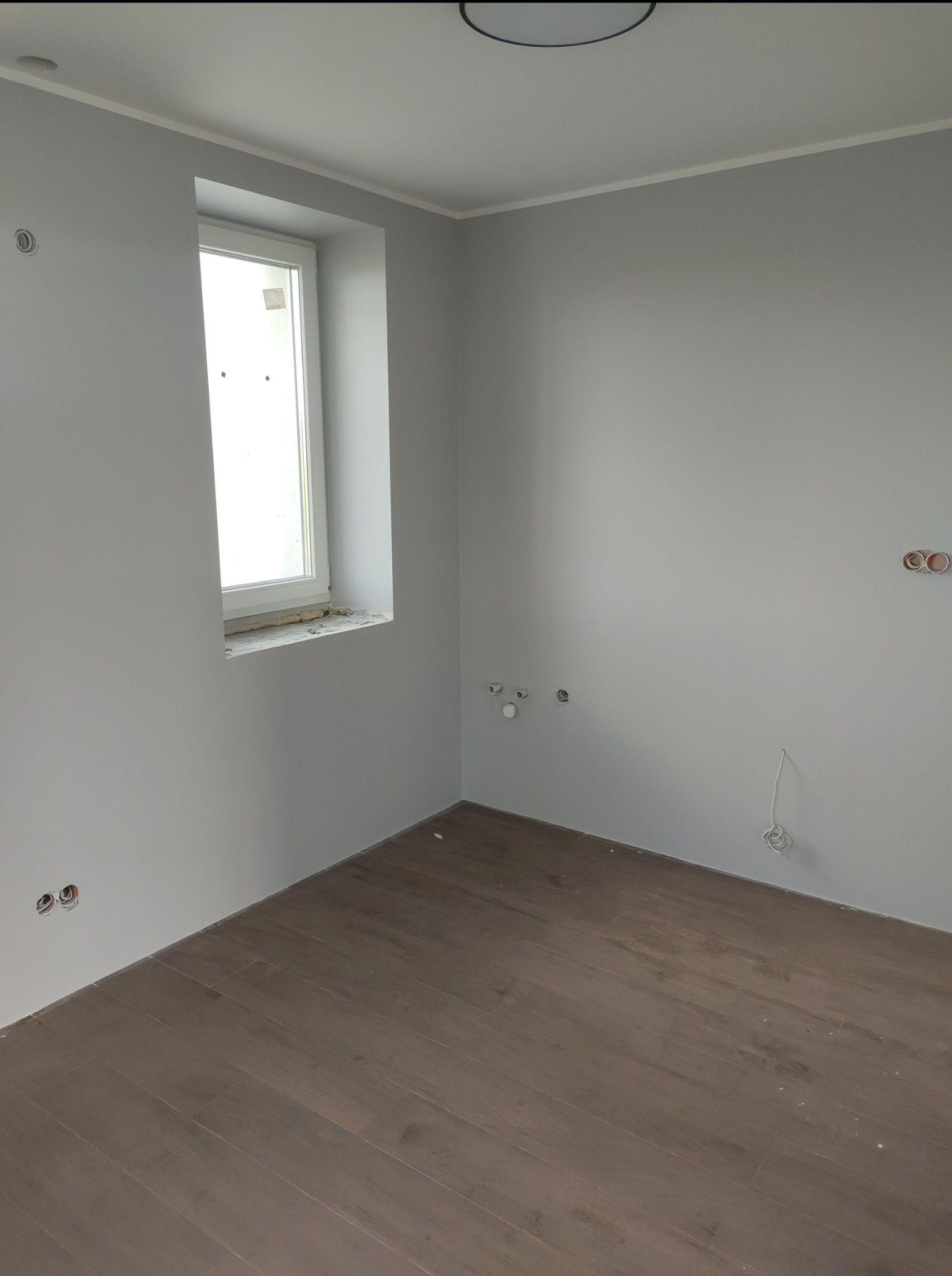 Pokój po odświeżeniu: szare ściany, brązowe panele podłogowe imitujące drewno, widoczne gniazdka elektryczne przed montażem osprzętu, okno z uszczelnieniem pianką.