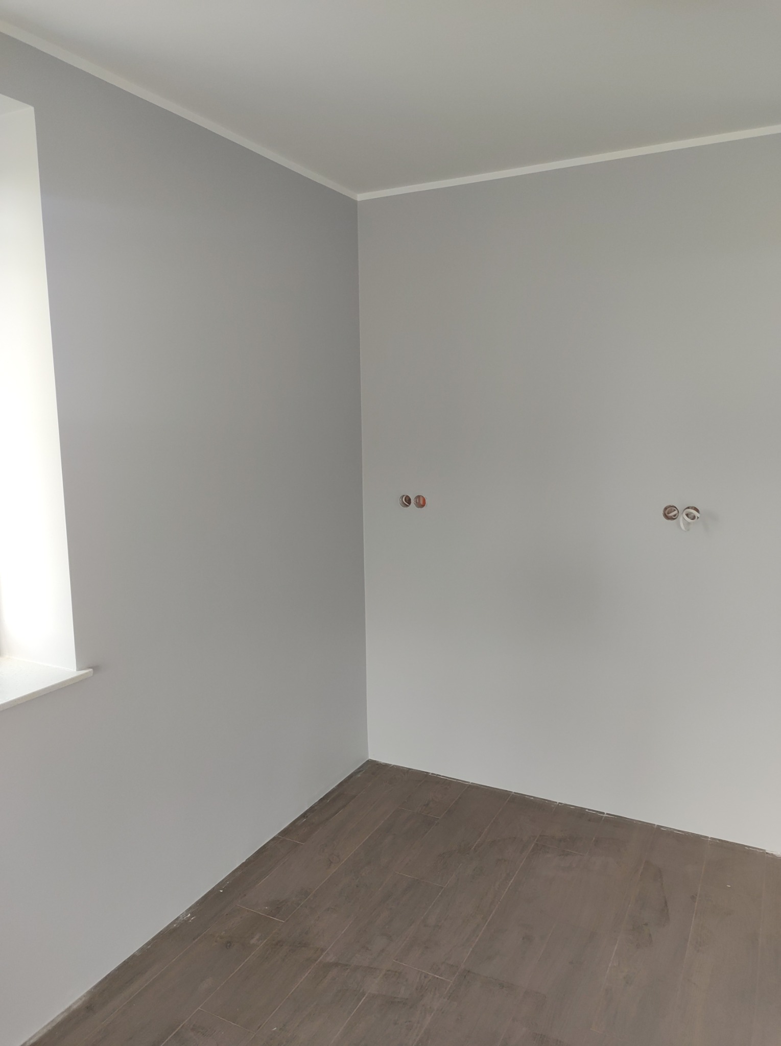 Pomalowane na szaro wnętrze z brązową podłogą z płytek imitujących drewno, widoczne puszki elektryczne na ścianie i biała listwa przypodłogowa.