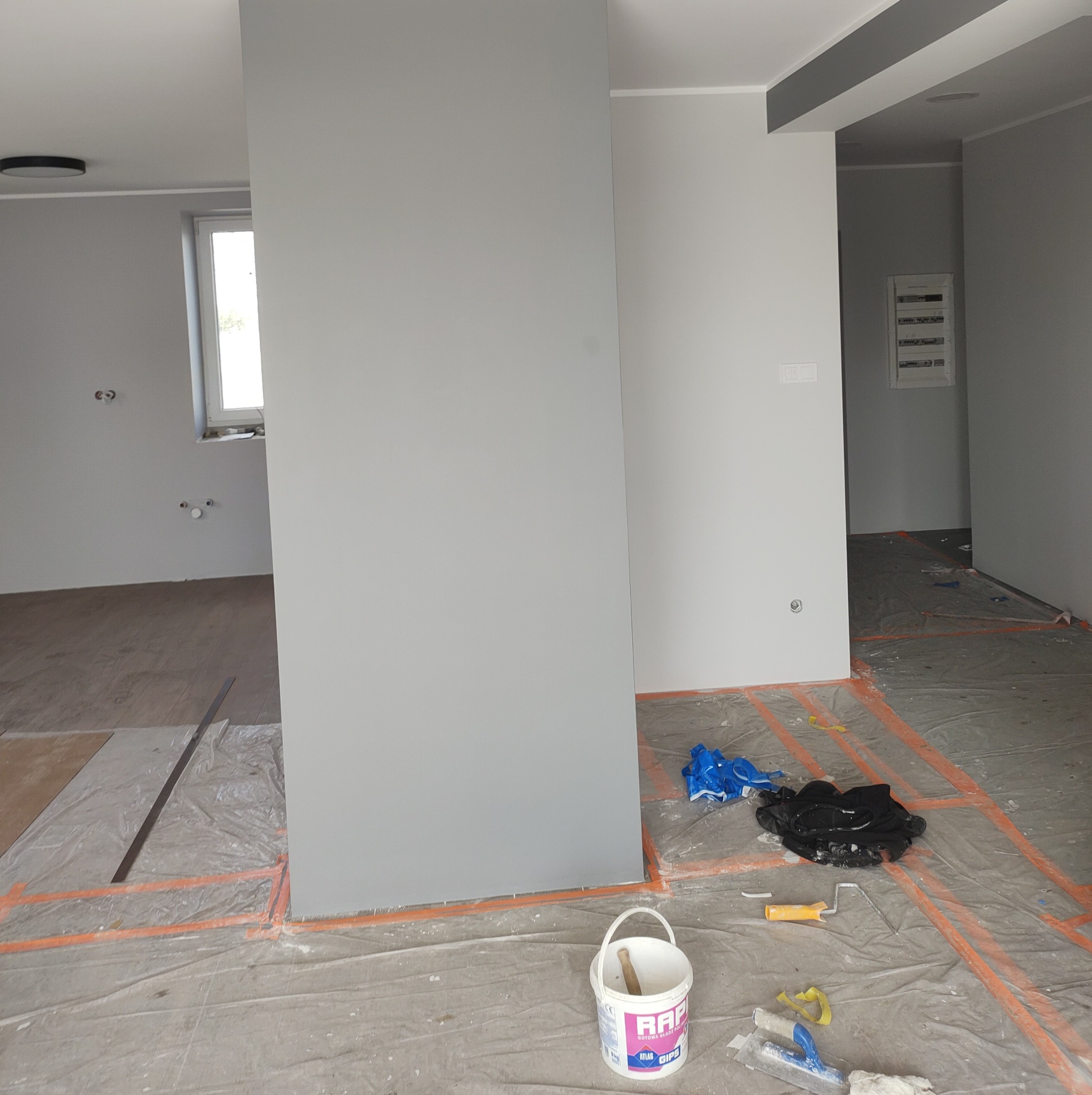 Szare ściany w trakcie malowania, podłoga zabezpieczona folią, widoczny kubeł z farbą Atlas Gips i narzędzia malarskie: wałek i szpachelka.