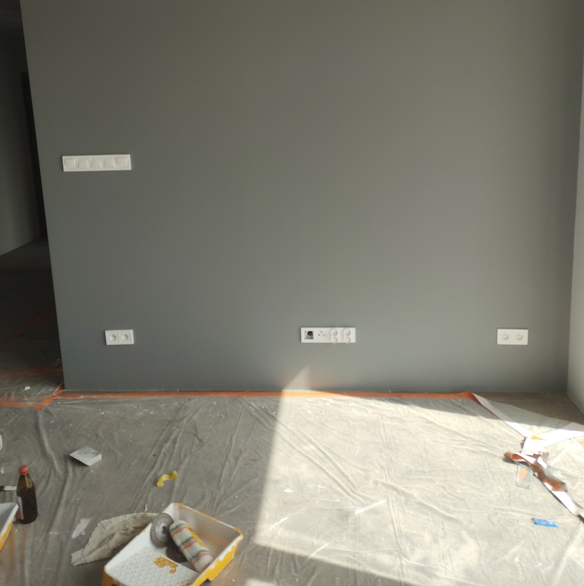 Świeżo pomalowana szara ściana z widocznymi gniazdkami elektrycznymi i włącznikiem światła, zabezpieczona folią malarską na podłodze, widoczne narzędzia malarskie w kuwecie.