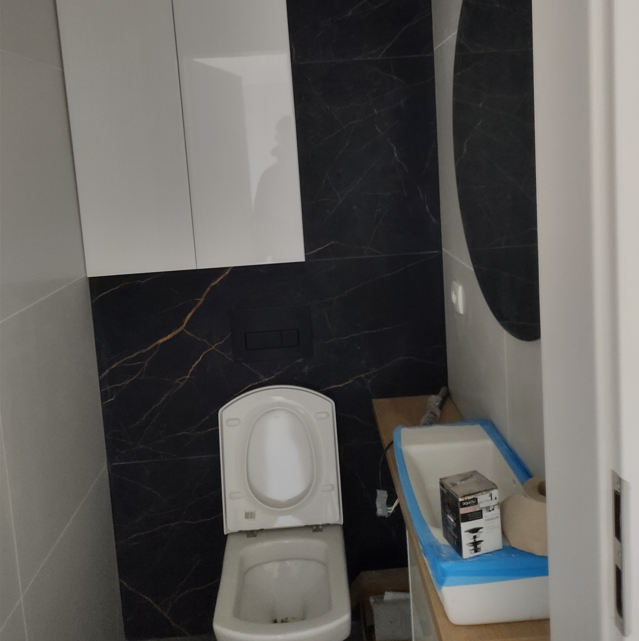 Nowoczesna łazienka w trakcie wykańczania: biała wisząca toaleta z otwartą klapą, czarna ściana z marmurowym wzorem, białe szafki i umywalka zabezpieczona folią malarską.