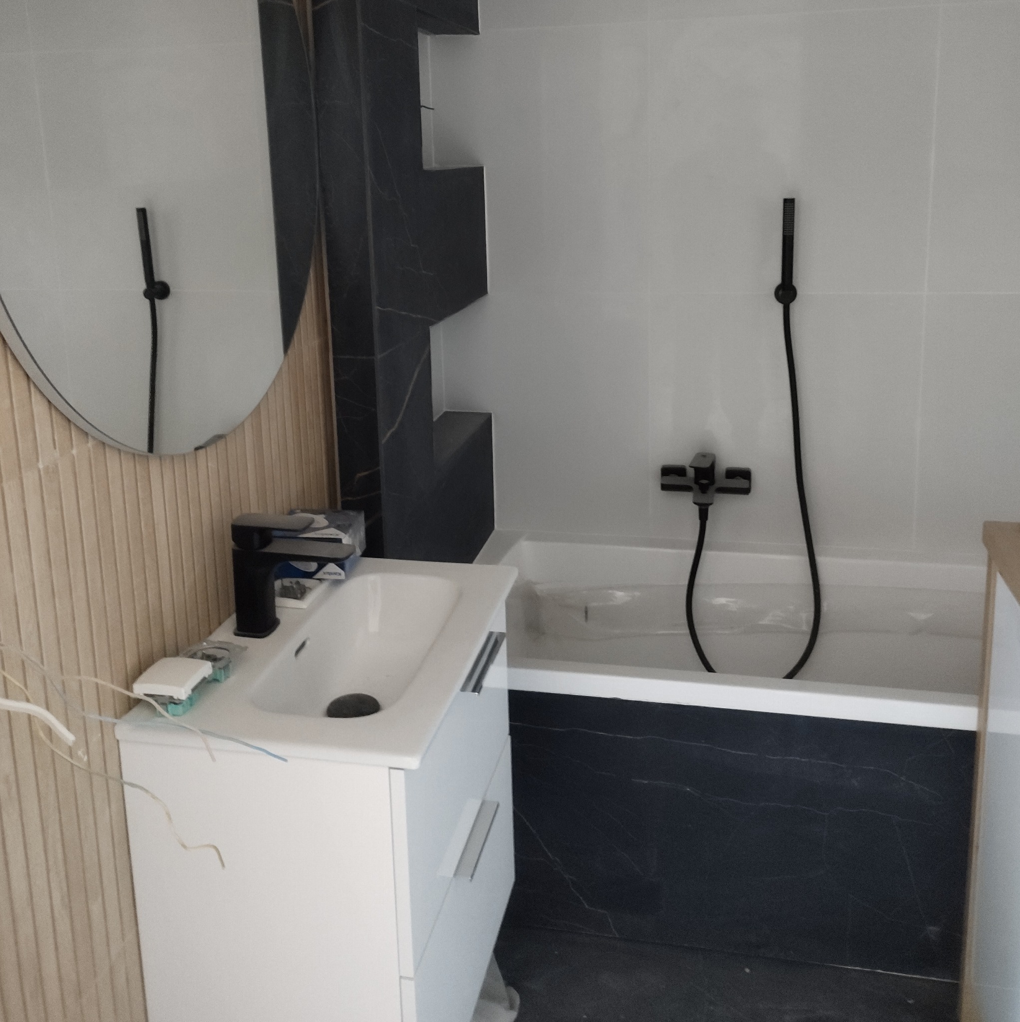 Nowoczesna łazienka z białą umywalką na szafce, czarną baterią, okrągłym lustrem z drewnianą ramą, wanną obudowaną czarnym marmurem i czarną armaturą.