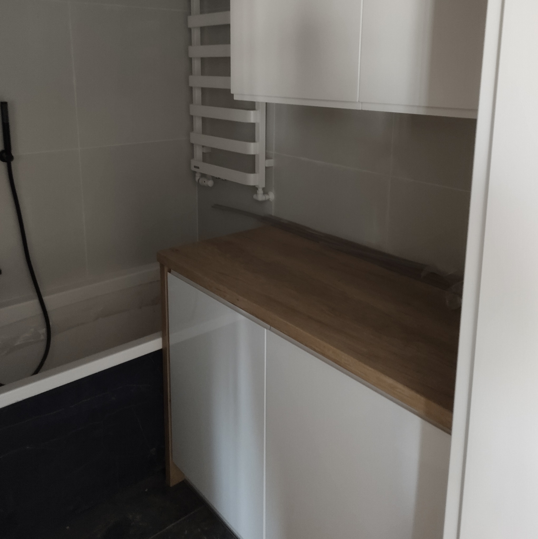 Nowoczesna łazienka z czarną podłogą, białymi płytkami i szafką pod umywalkę z drewnianym blatem, widoczny grzejnik drabinkowy.