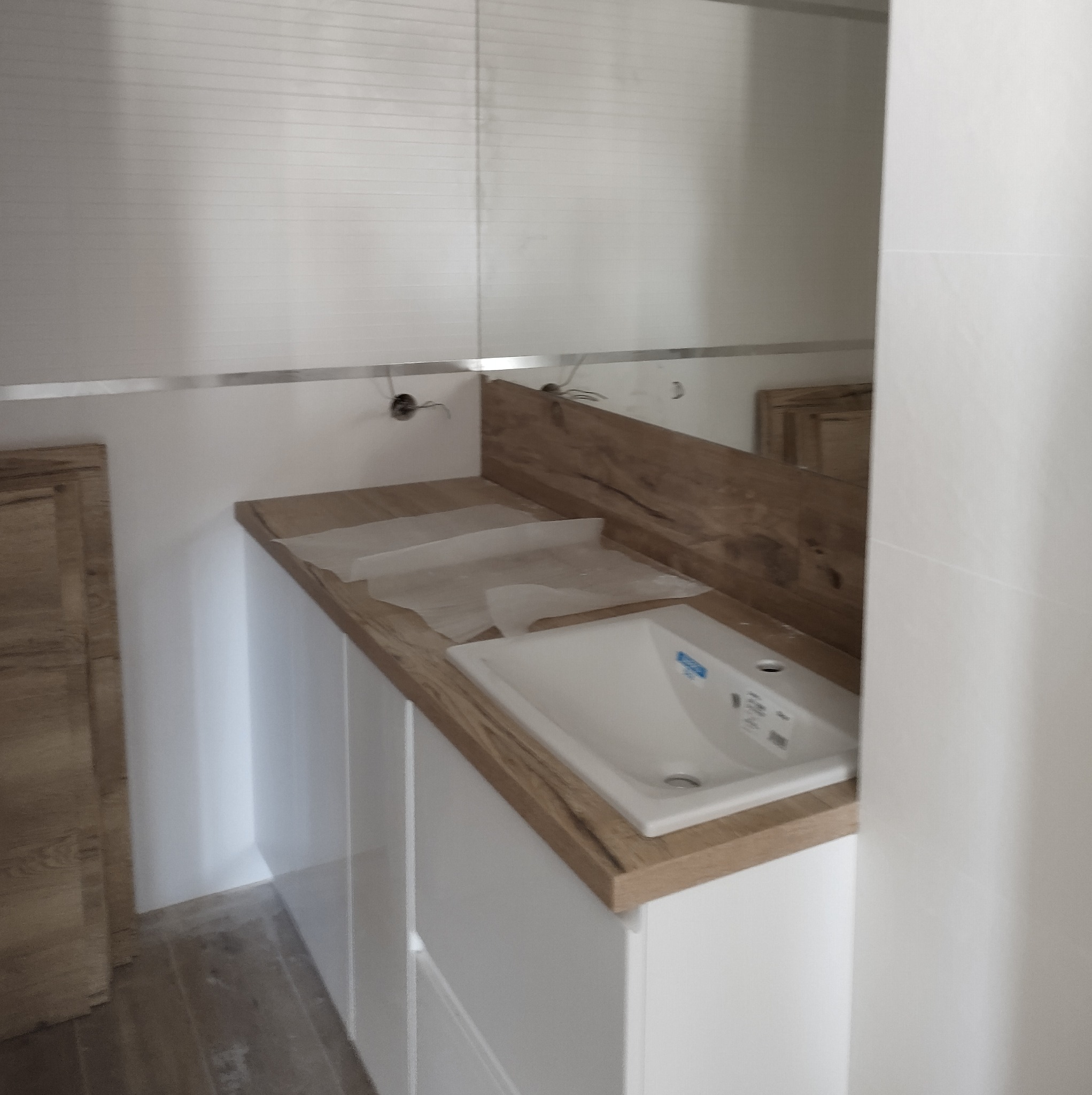 Biała umywalka wbudowana w szafkę z blatem imitującym drewno, zabezpieczona folią ochronną, na tle ściany z poziomymi panelami i lustra z widocznymi przewodami elektrycznymi.