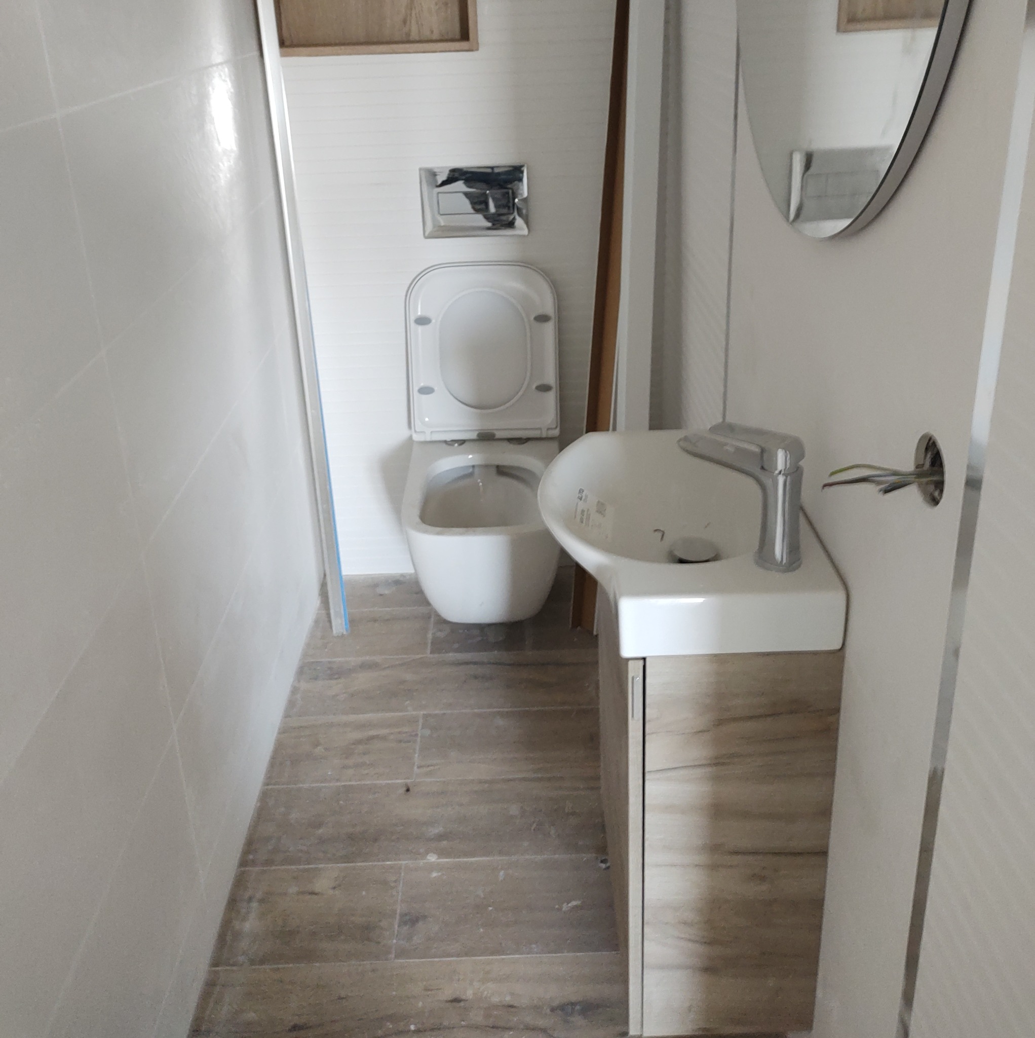 Wnętrze łazienki w trakcie wykańczania, widoczna podwieszana toaleta z otwartą klapą, umywalka z szafką w odcieniach drewna, białe płytki na ścianach i drewnopodobne panele na podłodze, wystające...