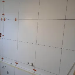 Białe płytki ścienne w trakcie montażu, z widocznymi krzyżykami dystansowymi i otworami na instalacje, ułożone na ścianie nad zabudową.