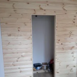 Ściana wykończona jasnymi, poziomymi panelami drewnianymi z widocznymi sękami, z otworem drzwiowym prowadzącym do pomieszczenia z widocznymi narzędziami i materiałami budowlanymi.