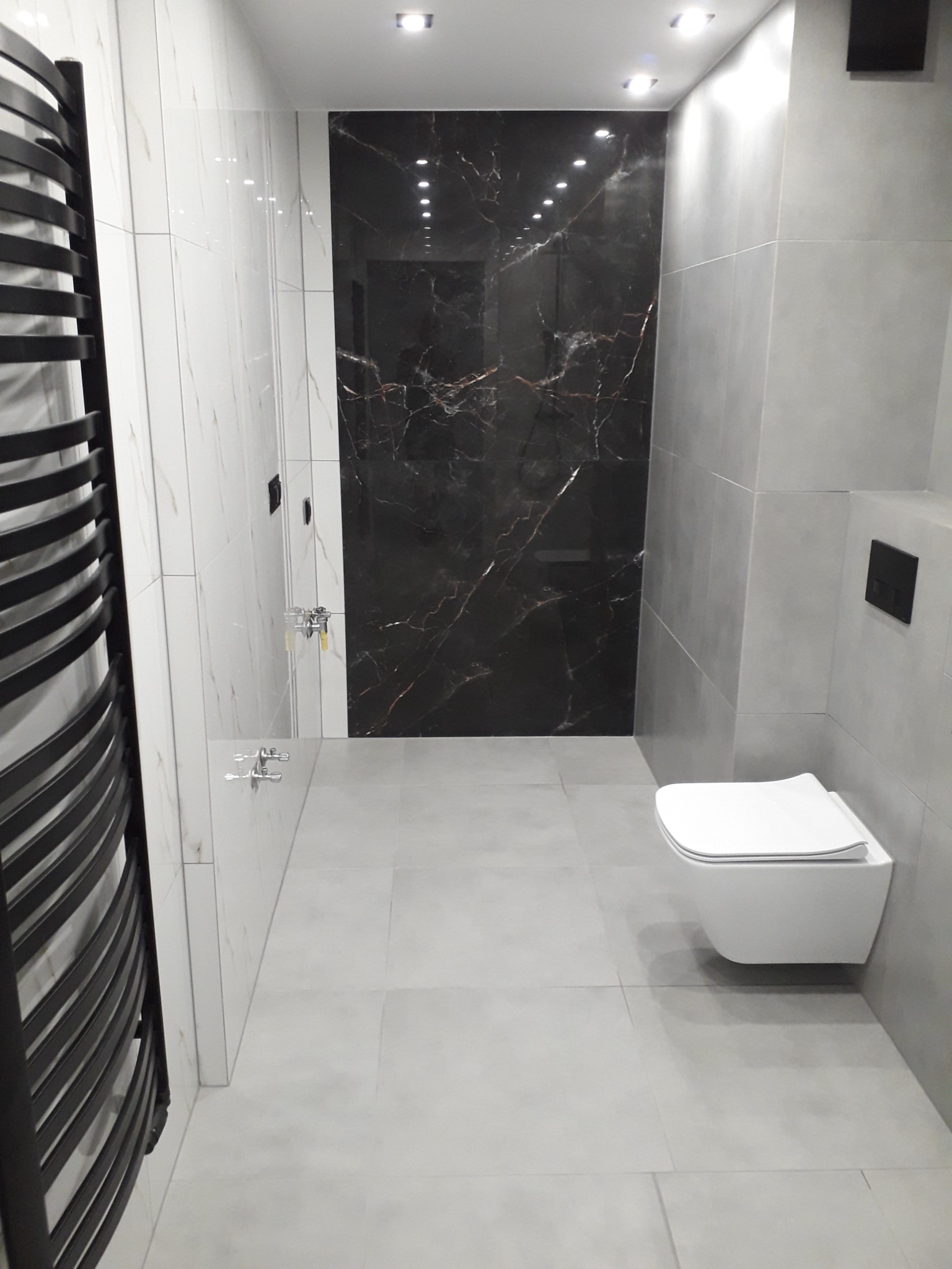 Nowoczesna łazienka z szarymi płytkami podłogowymi i ściennymi, czarną ścianą z marmurowym wzorem, wiszącą toaletą oraz czarnym grzejnikiem drabinkowym po lewej stronie.