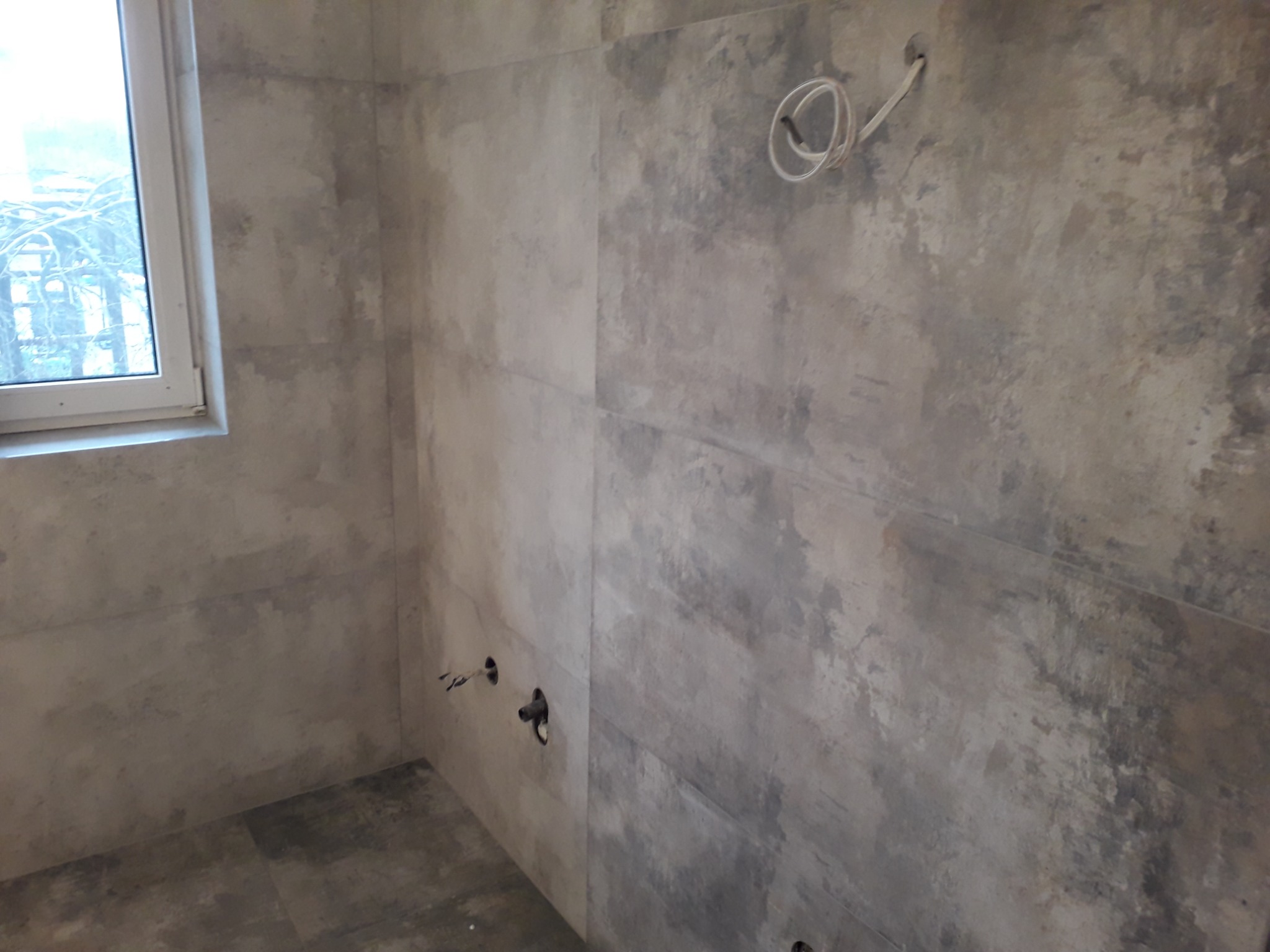 Surowa łazienka w trakcie wykończenia z szarymi płytkami imitującymi beton, widoczne wystające kable i rury, okno w lewym górnym rogu.