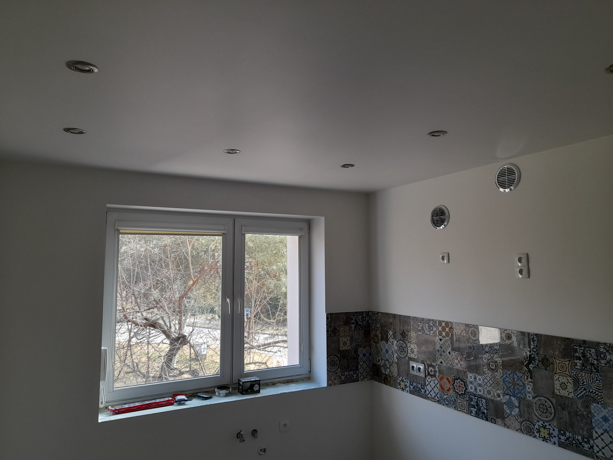 Świeżo wykończona łazienka z patchworkowymi płytkami nad blatem, widoczne otwory wentylacyjne i wpuszczone oświetlenie sufitowe, okno z widokiem na drzewo bez liści.