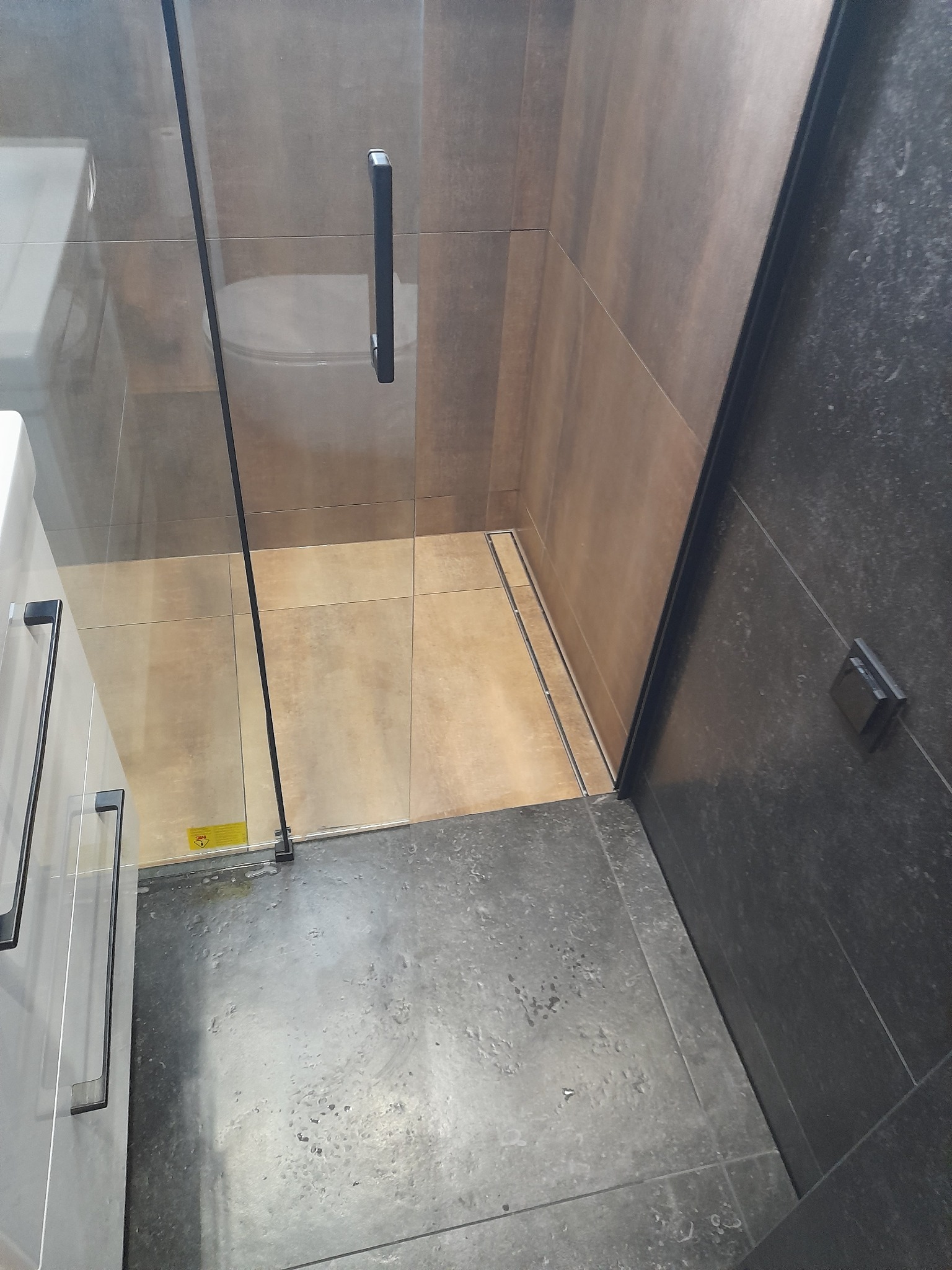 Nowoczesna kabina prysznicowa z czarnymi detalami, szklane drzwi z minimalistycznym uchwytem, widoczny odpływ liniowy i mieszanka płytek w odcieniach brązu i szarości.