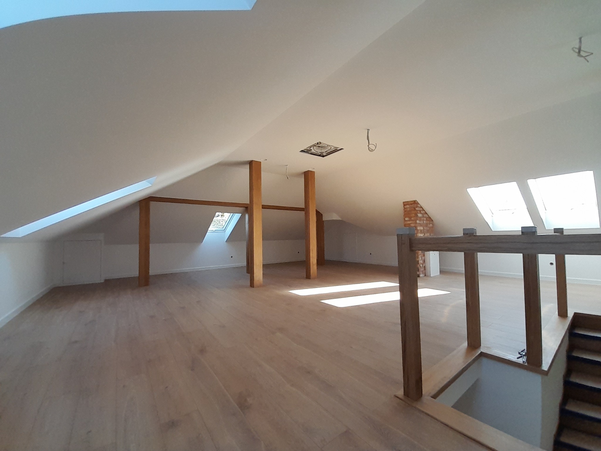 Przestronne poddasze po remoncie z drewnianymi słupami, jasną podłogą i oknami dachowymi wpuszczającymi dużo światła, widoczny fragment schodów z drewnianą balustradą.