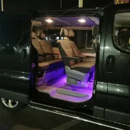 Wnętrze czarnego busa z otwartymi drzwiami, widoczne szare fotele z podłokietnikami i nastrojowe, fioletowe oświetlenie pod siedzeniami.