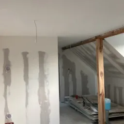 Poddasze w trakcie prac wykończeniowych: widoczne płyty karton-gips na ścianach, szpachlowanie w pionowe pasy, drewniana konstrukcja stropu, przewody elektryczne wystające ze ściany i sterta płyt...