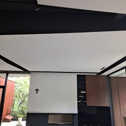 DOBRO-BUD OLEH LANOVENKO - Nowoczesne wnętrze z białym sufitem podwieszanym z czarnymi listwami, białą ścianą z kominkiem i czarnym krzyżem, widok na ogród przez okno.