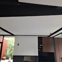 Nowoczesne wnętrze z białym sufitem podwieszanym z czarnymi listwami, białą ścianą z kominkiem i czarnym krzyżem, widok na ogród przez okno.