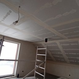 DOBRO-BUD OLEH LANOVENKO - Poddasze w trakcie wykańczania, widoczne płyty karton-gips na skosach i suficie, łączenia szpachlowane, drabina aluminiowa, surowa żarówka zwisająca z sufitu, okno w białej ramie.