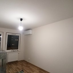 DOBRO-BUD OLEH LANOVENKO - Wnętrze pokoju po odświeżeniu: świeżo pomalowane ściany w odcieniu bieli, drewnopodobna podłoga, okna z widokiem na nocne miasto, minimalistyczna lampa sufitowa i klimatyzator na ścianie.