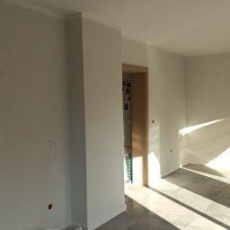 DOBRO-BUD OLEH LANOVENKO - Jasne, świeżo pomalowane wnętrze z szarymi płytkami podłogowymi, widoczne wiadro farby Flügger Flutex 2S przy ścianie i wpadające przez okno promienie słoneczne.