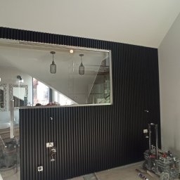 DOBRO-BUD OLEH LANOVENKO - Montaż czarnych paneli ściennych z pionowymi listwami w pomieszczeniu z widocznym fragmentem łazienki za szklaną przegrodą, widoczne kable elektryczne wystające ze ściany, drabina i narzędzia.