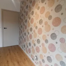 Pokój po remoncie z nową tapetą w okrągłe wzory w odcieniach beżu, brązu i szarości oraz jasną drewnianą podłogą i białymi listwami przypodłogowymi, widoczna biała ściana i drzwi.