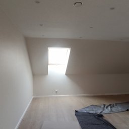 DOBRO-BUD OLEH LANOVENKO - Jasne poddasze po remoncie: świeżo pomalowane ściany w odcieniach bieli, panele podłogowe, okno dachowe wpuszczające światło, widoczna kolumna nośna.