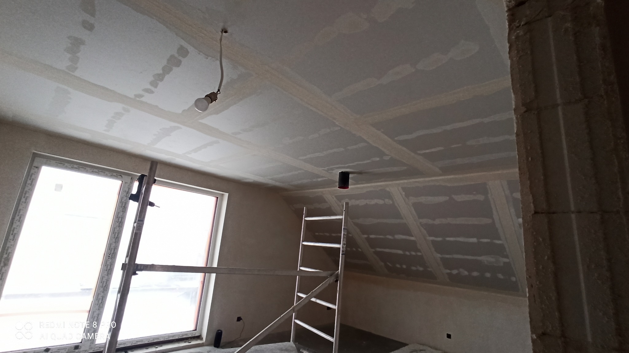 Poddasze w trakcie wykańczania, widoczne płyty karton-gips na skosach i suficie, łączenia szpachlowane, drabina aluminiowa, surowa żarówka zwisająca z sufitu, okno w białej ramie.