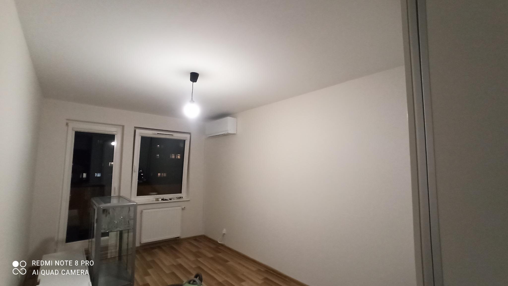 Wnętrze pokoju po odświeżeniu: świeżo pomalowane ściany w odcieniu bieli, drewnopodobna podłoga, okna z widokiem na nocne miasto, minimalistyczna lampa sufitowa i klimatyzator na ścianie.