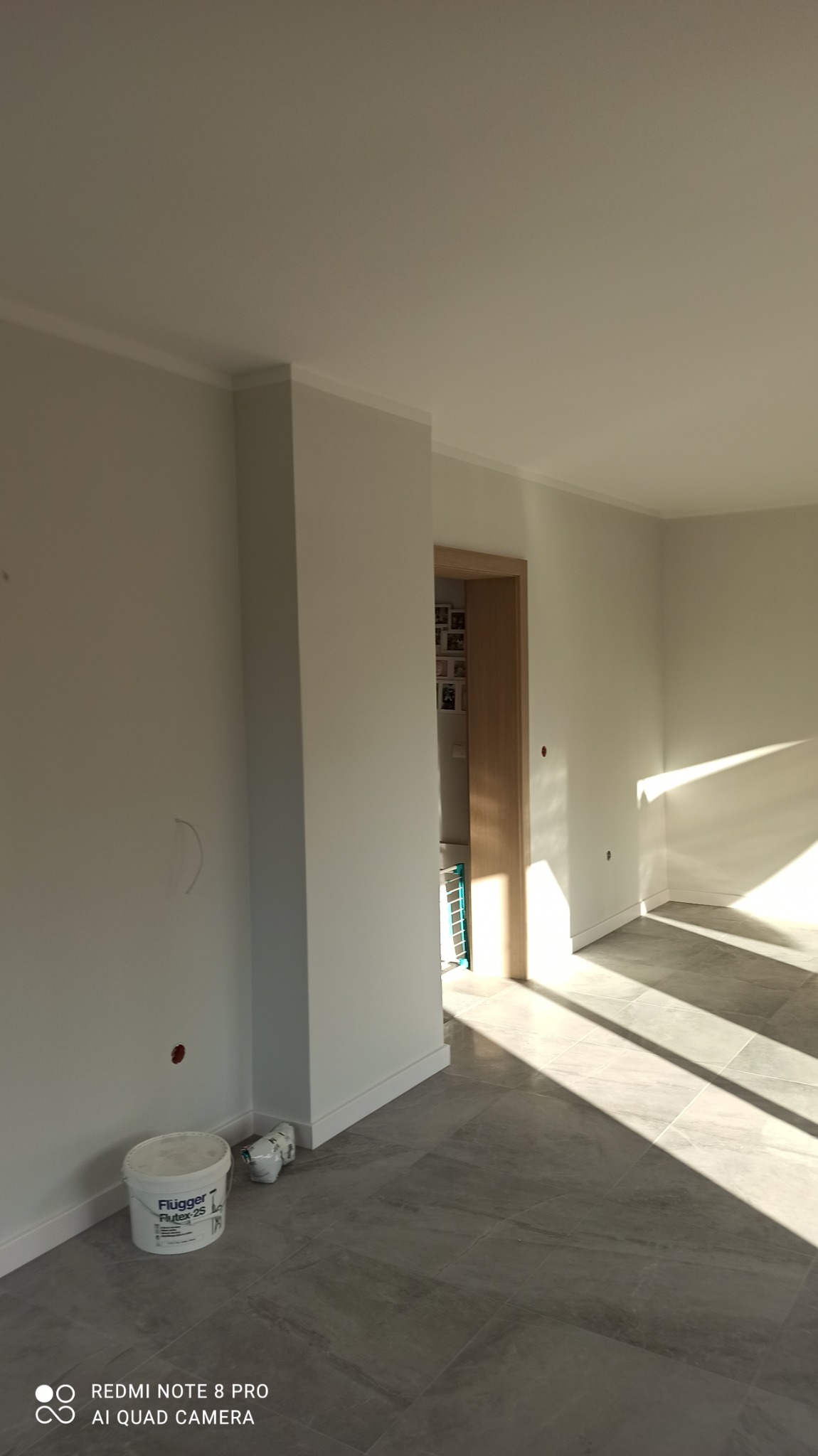 Jasne, świeżo pomalowane wnętrze z szarymi płytkami podłogowymi, widoczne wiadro farby Flügger Flutex 2S przy ścianie i wpadające przez okno promienie słoneczne.