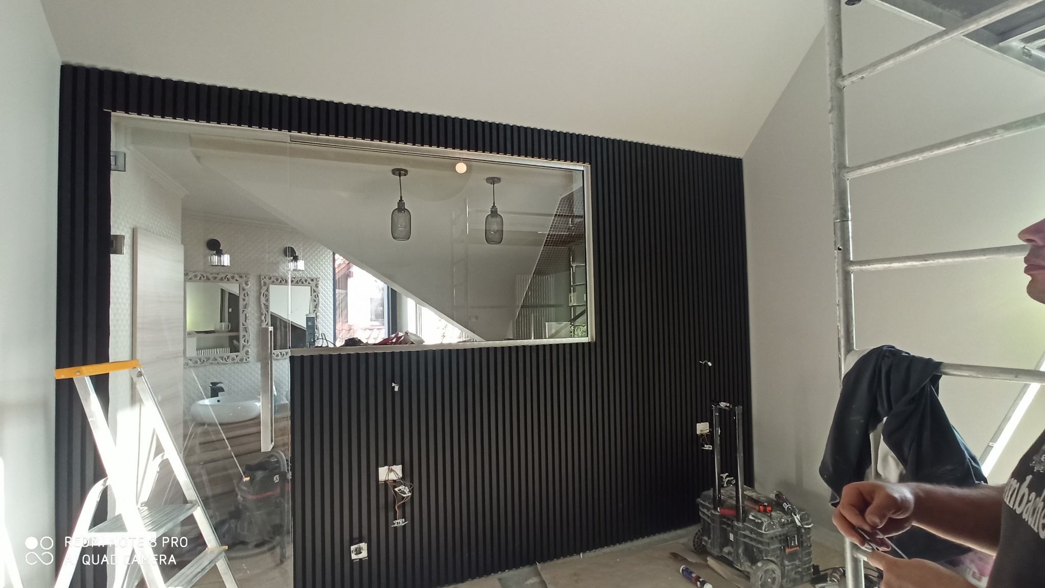 Montaż czarnych paneli ściennych z pionowymi listwami w pomieszczeniu z widocznym fragmentem łazienki za szklaną przegrodą, widoczne kable elektryczne wystające ze ściany, drabina i narzędzia.