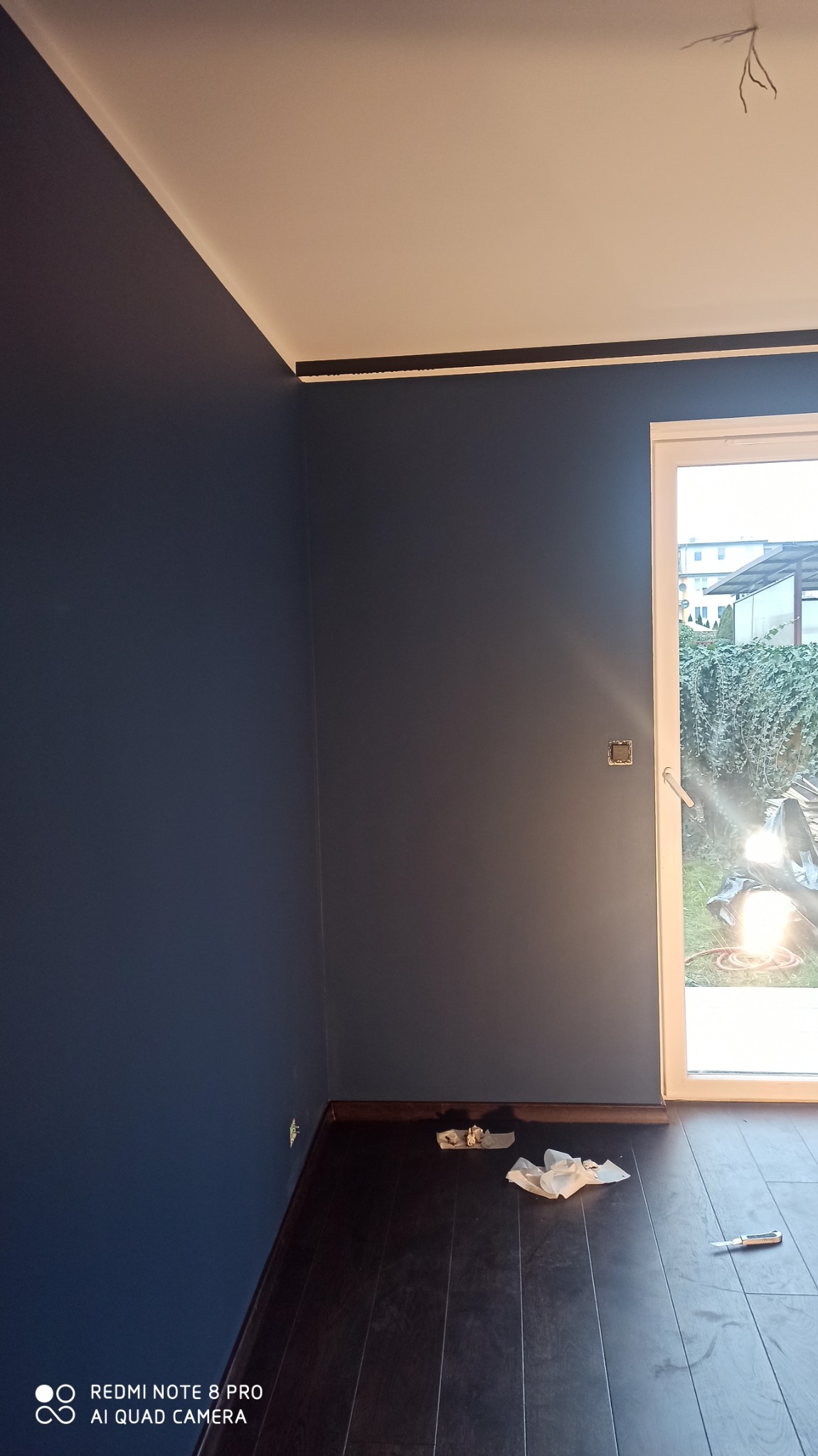 Pomieszczenie po malowaniu ścian na ciemny niebieski kolor, widoczne listwy przypodłogowe i sufitowe, okno z widokiem na ogród, na podłodze resztki po malowaniu i nóż do tapet.