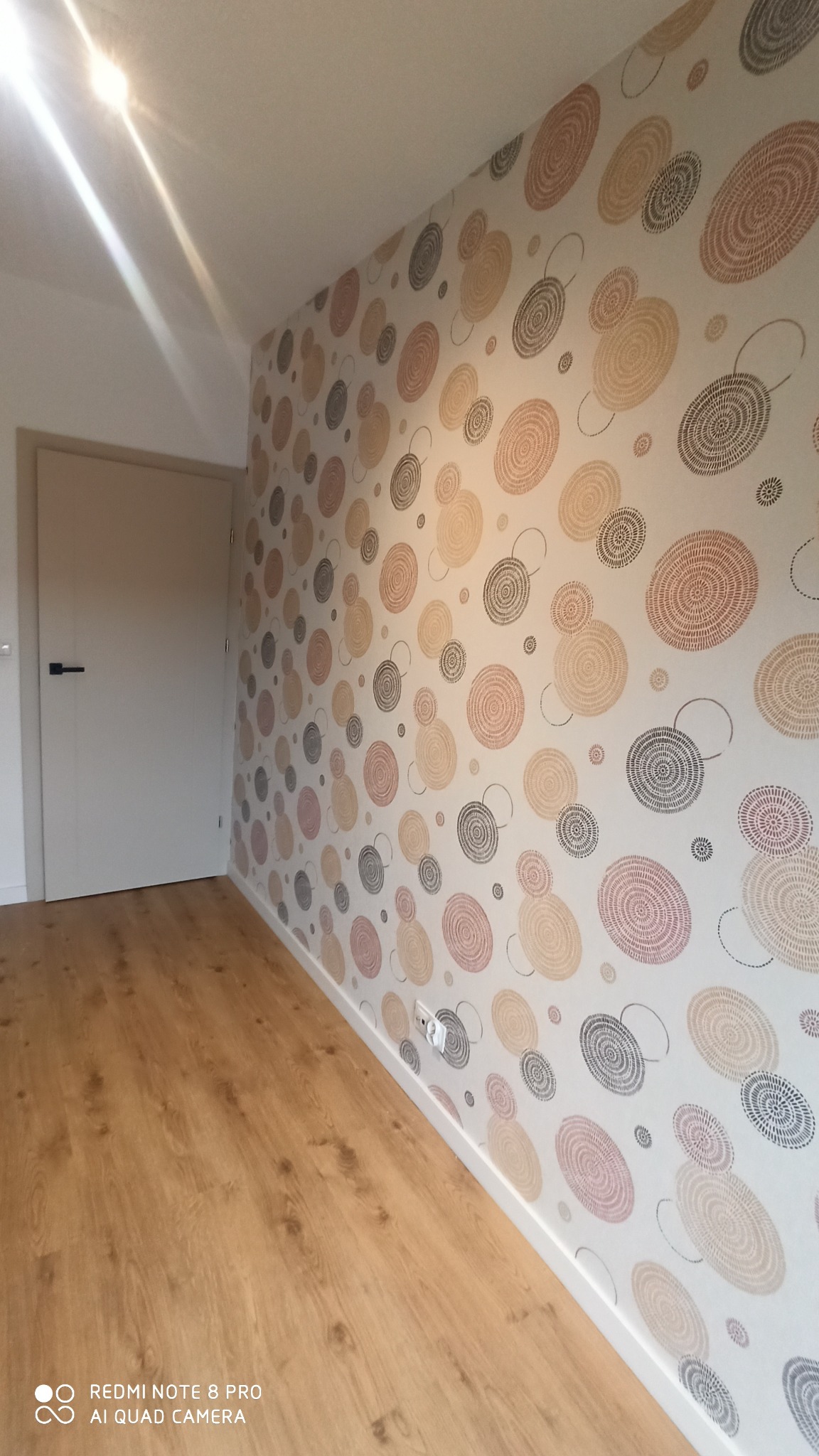 Pokój po remoncie z nową tapetą w okrągłe wzory w odcieniach beżu, brązu i szarości oraz jasną drewnianą podłogą i białymi listwami przypodłogowymi, widoczna biała ściana i drzwi.