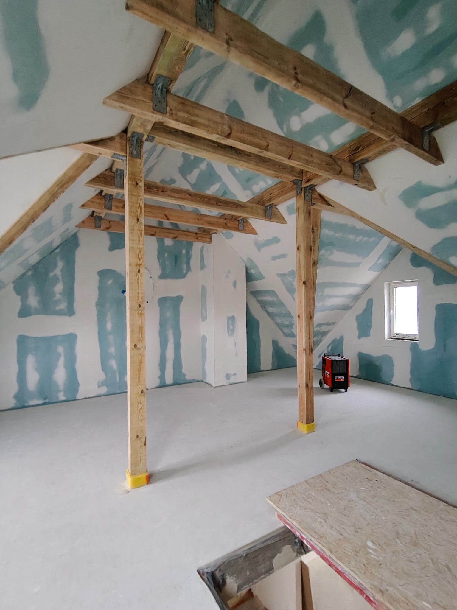 Poddasze w trakcie prac wykończeniowych: surowe drewniane belki, ściany z widocznymi łączeniami płyt, jasna podłoga. W tle nagrzewnica.