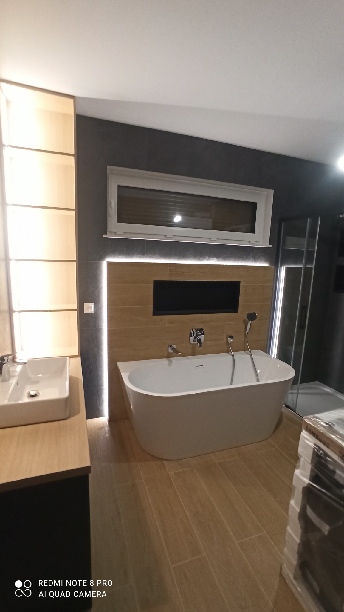 Nowoczesna łazienka z wolnostojącą wanną, drewnopodobnymi płytkami i oświetleniem LED. Umywalka nablatowa i kabina prysznicowa w tle. Minimalistyczny design.