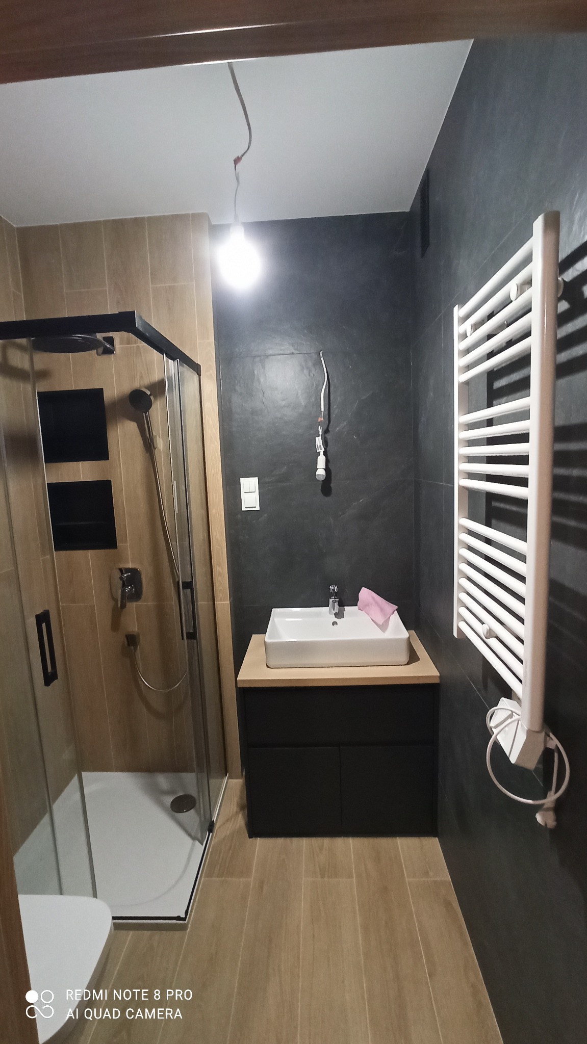 Nowoczesna łazienka z kabiną prysznicową, umywalką na szafce i grzejnikiem drabinkowym. Kontrastujące płytki: drewnopodobne i grafitowe. Widoczne niedokończone podłączenie oświetlenia.