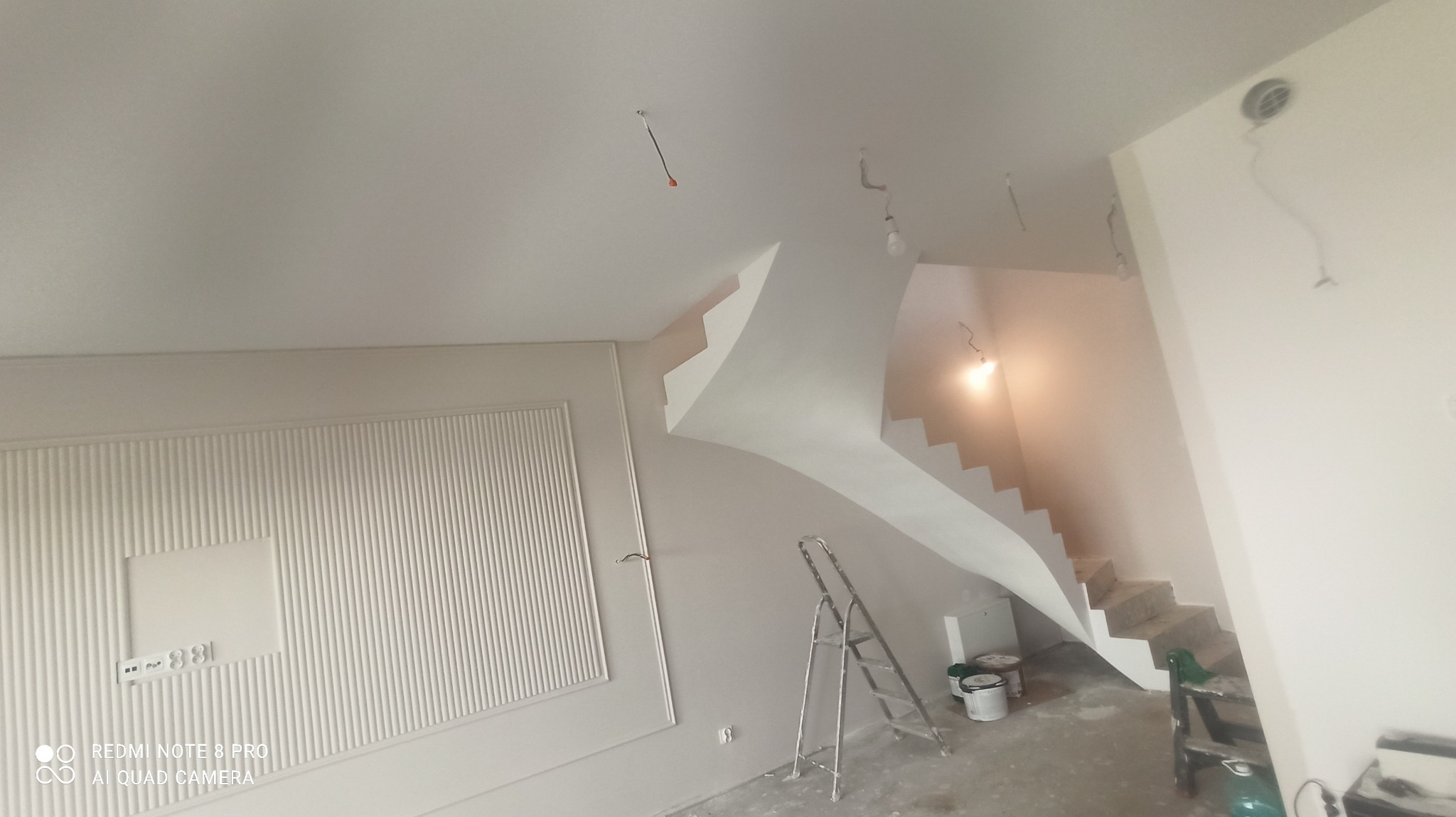 Jasne wnętrze po malowaniu: ściany w odcieniach bieli, dekoracyjna sztukateria, schody z widocznym oświetleniem, drabina malarska i akcesoria malarskie.