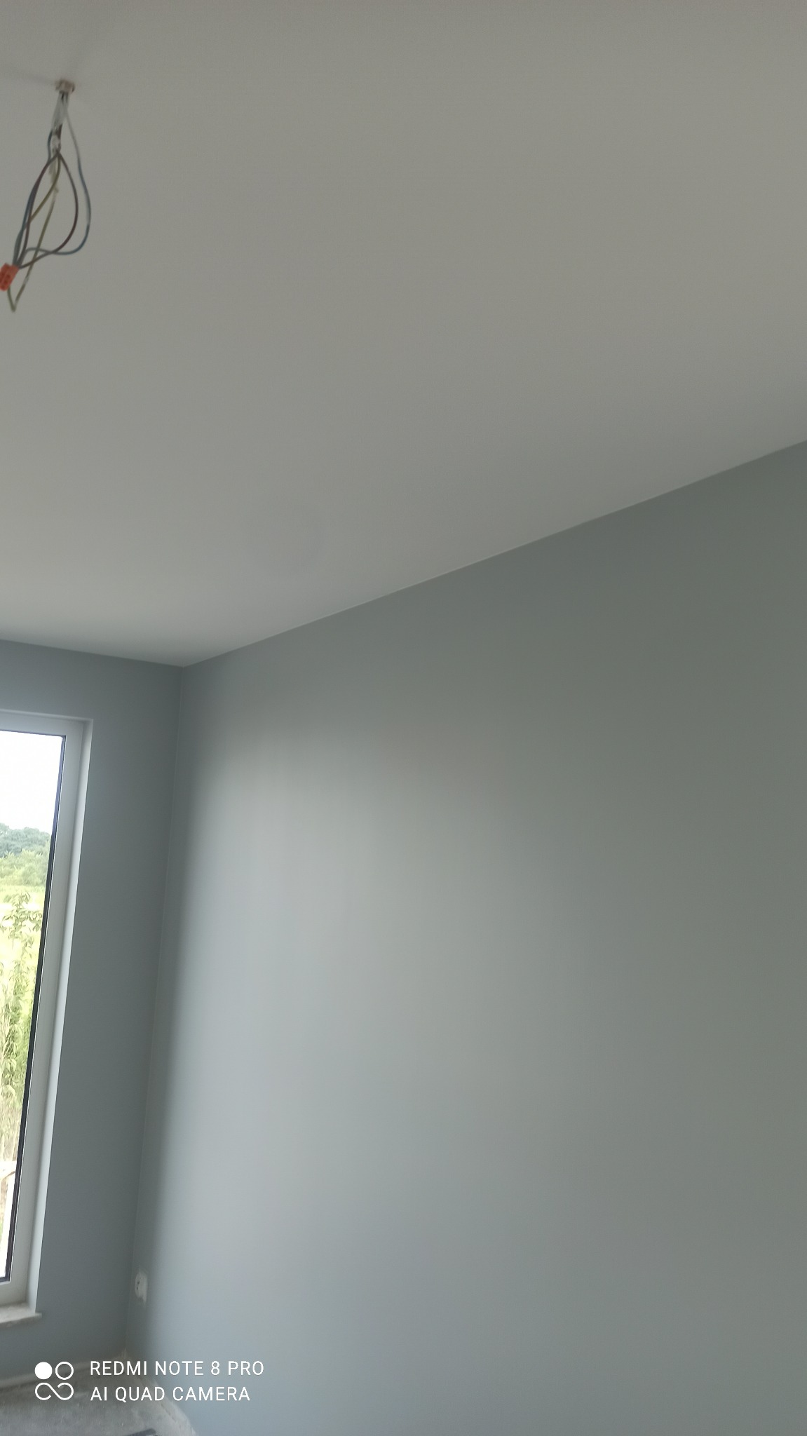 Pomieszczenie po malowaniu ścian na szaro, widoczne okno i wystające kable z sufitu. Świeżo pomalowane ściany w odcieniach szarości, sufit biały.