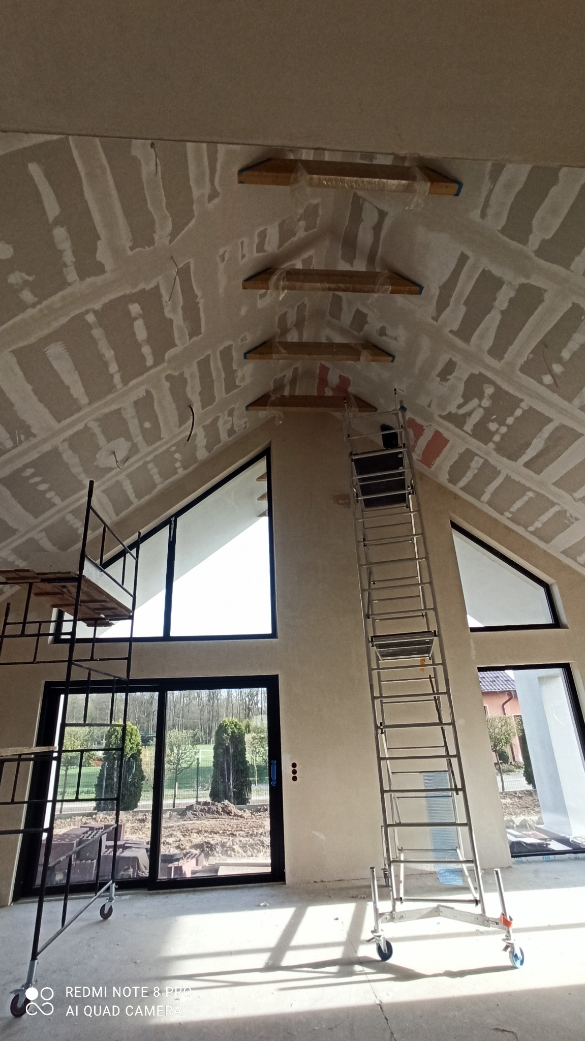 Skośny sufit poddasza po szpachlowaniu, widoczne łączenia płyt gipsowych, rusztowanie i drabina. Duże okna z widokiem na zewnątrz. Etap prac wykończeniowych.