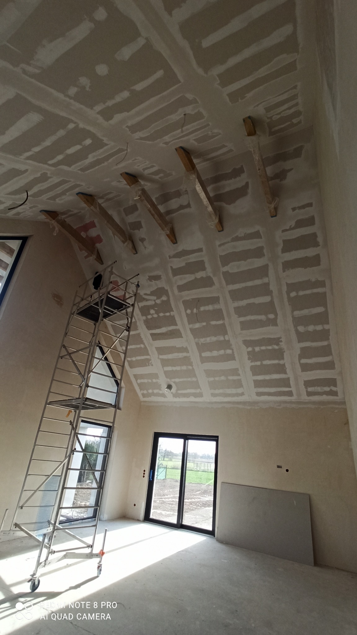 Skośny sufit poddasza po szpachlowaniu, widoczne drewniane belki, rusztowanie w pomieszczeniu z oknami. Prace wykończeniowe w jasnym wnętrzu.