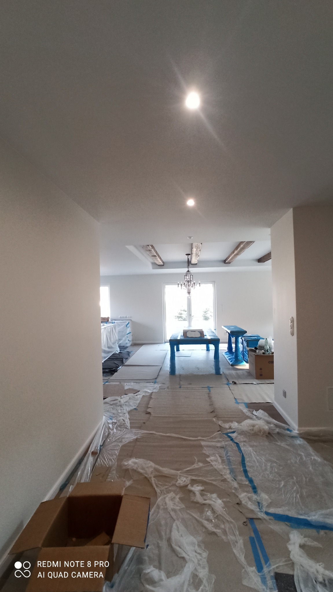 Wnętrze w trakcie prac wykończeniowych: zabezpieczona podłoga folią, świeżo pomalowane ściany, widoczny fragment sufitu z dekoracyjnymi belkami i żyrandolem.