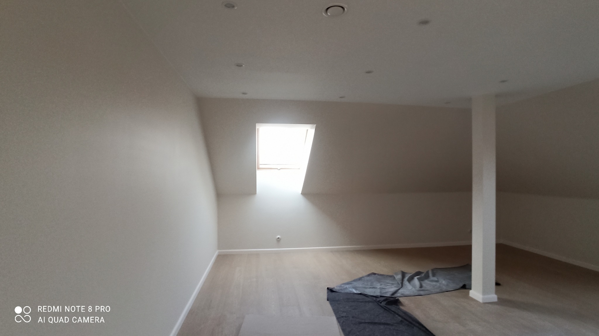 Jasne poddasze po remoncie: świeżo pomalowane ściany w odcieniach bieli, panele podłogowe, okno dachowe wpuszczające światło, widoczna kolumna nośna.